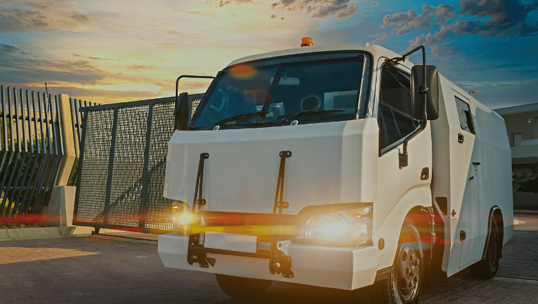 A G4S security armored van with its lights on, parked in front of a fenced industrial facility at sunset.