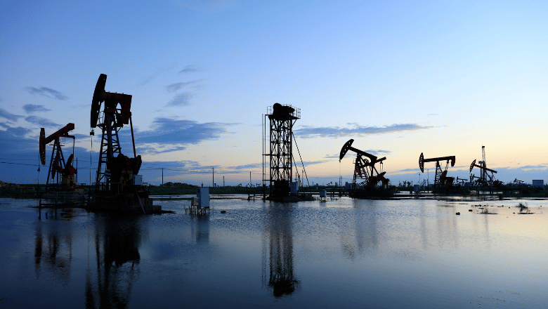 Silhouette of multiple oil pump jacks (nodding donkeys) and industrial drilling rigs operating in a shallow body of water at dusk or dawn.