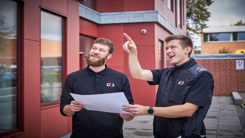 Two individuals wearing black G4S uniforms stand outside a building with red walls and large windows.