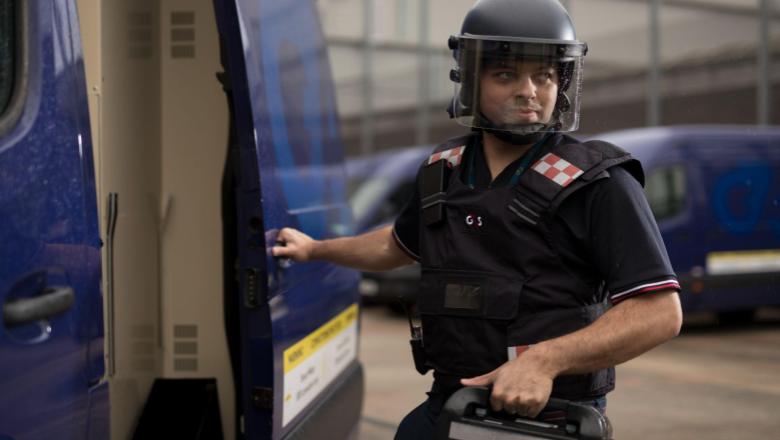 A G4S security guard in body armour and a helmet loading a secure case into a blue armoured van.