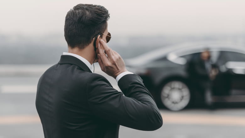 Stock image of a close protection officer touching their earpiece as a passenger exits a parked car