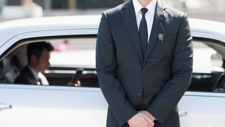 A stock image of a close protection officer guarding a vehicle. A man is reading his mobile phone in the back seats of the vehicle.