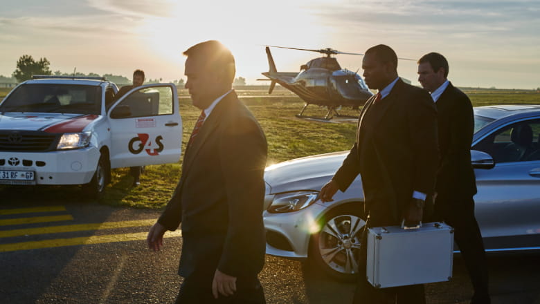 Two Close Protection Officers (CPOs) escorting an executive businessman off an airport tarmac. There is a G4S vehicle and non-branded helicopter in the background