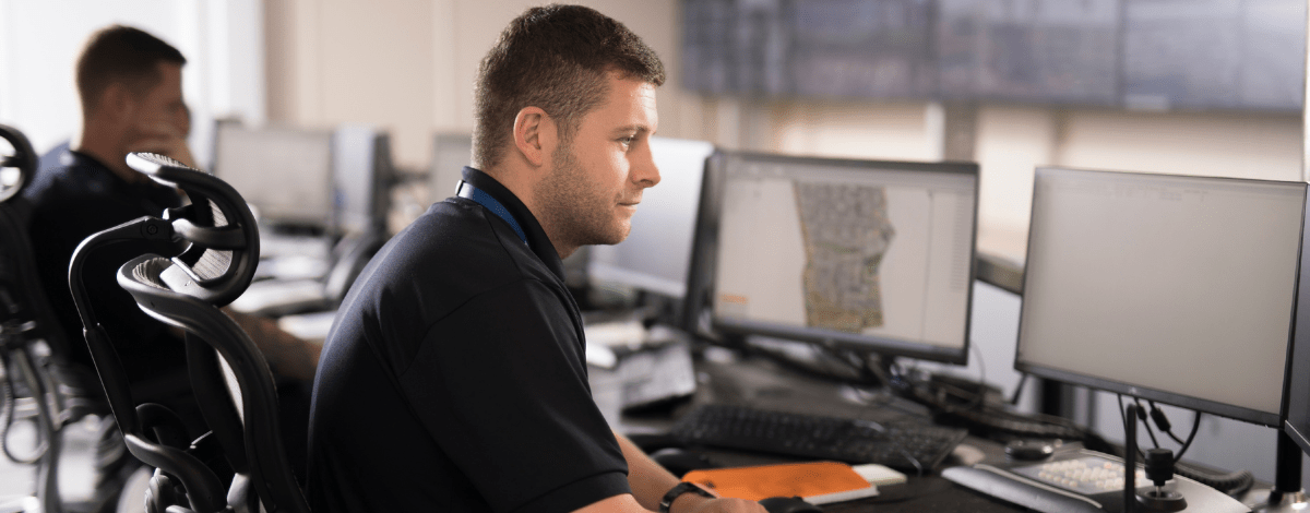 A man in a dark polo shirt working at a professional workstation with multiple computer monitors in a bright operations centre