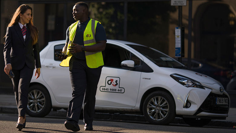 A lady in a suit walking on the street and interacting with a male G4S employee in a yellow vest