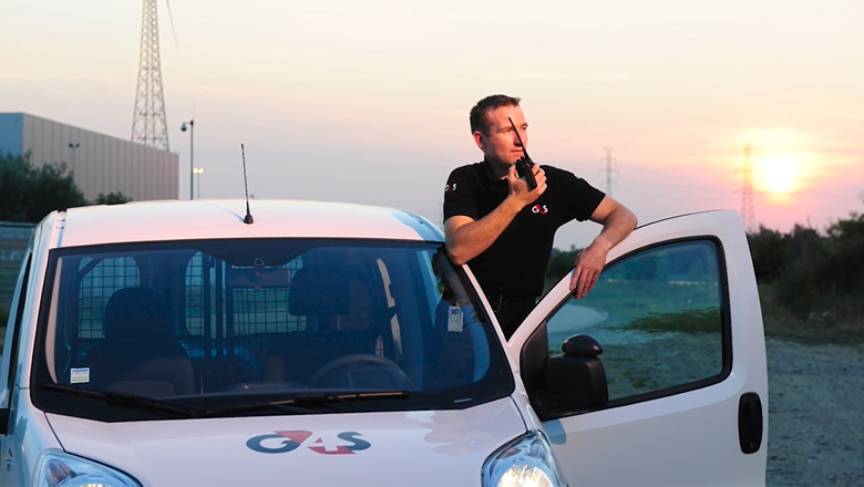 A man in G4S uniform holding a machine and leaning towards a white car