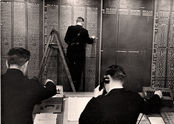A man writing on a blackboard and 2 receptionists answering phone calls behind in 1955