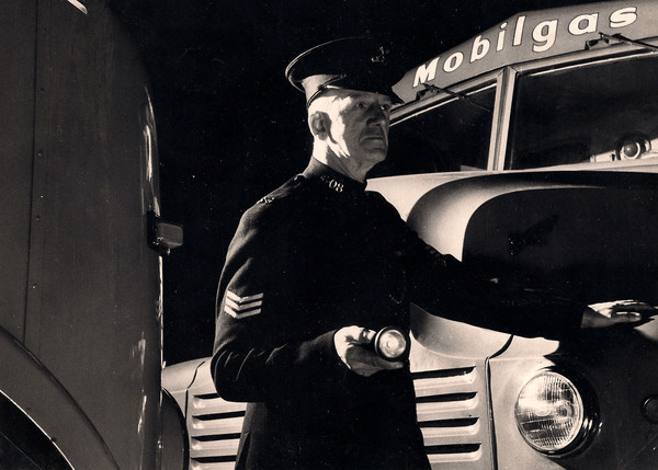 A security guard holding a flashlight standing in front of a car with Mobilgas above the windscreen