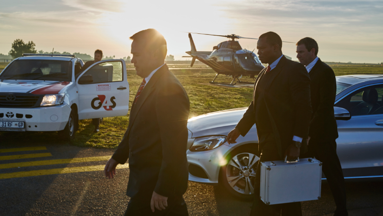 Two Close Protection Officers (CPOs) escorting an executive businessman off an airport tarmac. There is a G4S vehicle and non-branded helicopter in the background