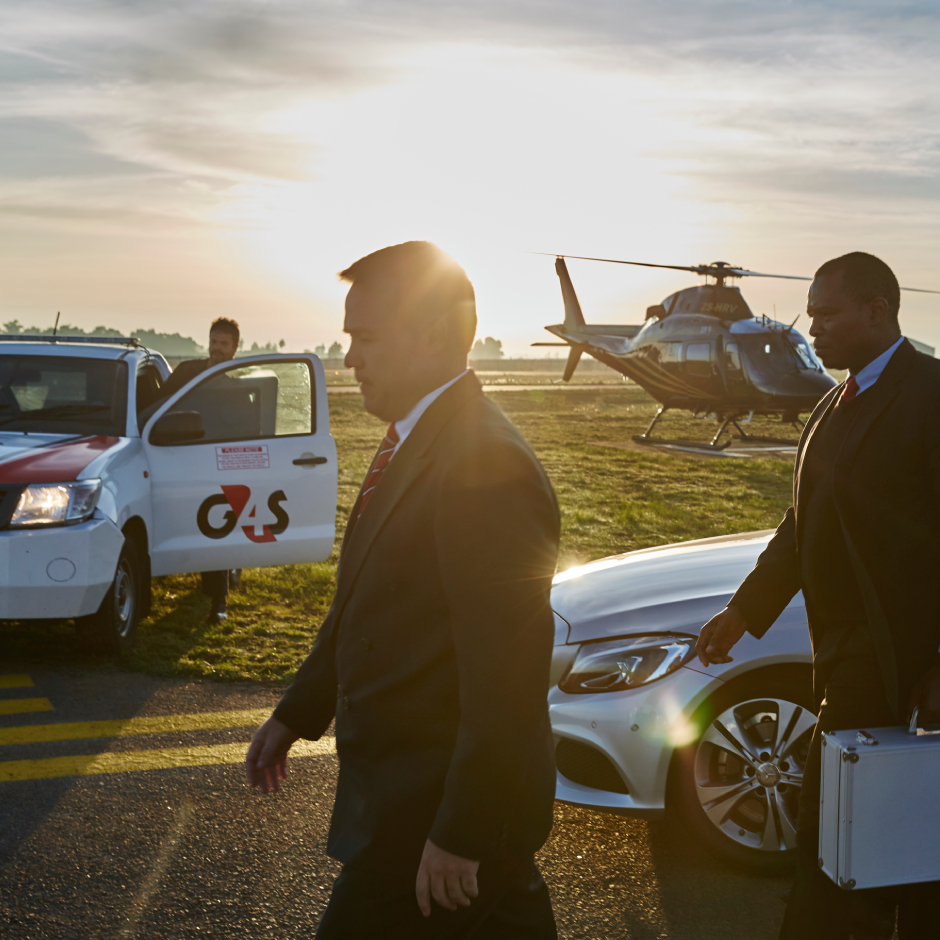 A Close Protection Officers (CPOs) escorting an executive businessman off an airport tarmac. There is a G4S vehicle and non-branded helicopter in the background