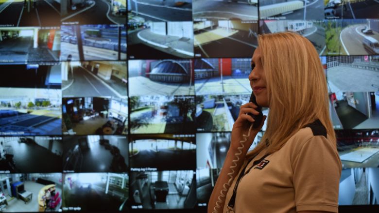 A lady answering phone calls standing in front of a wall of CCTV monitor screens